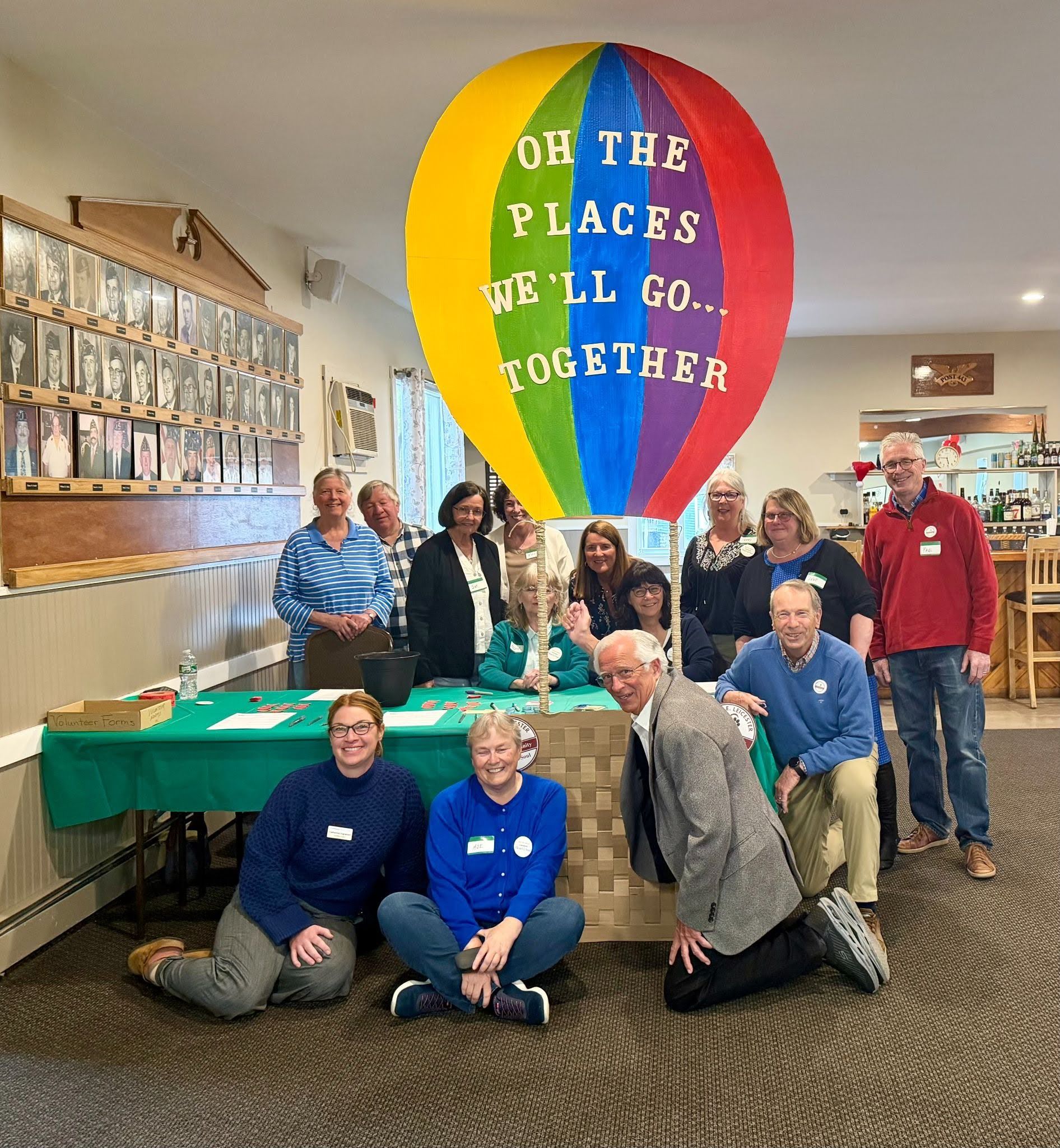 A group of Leicester residents posing in front of a paper banner in the shape of a rainbow hot air ballon with the text "Oh the places we'll go...together."